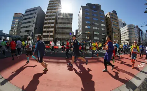 Atleti corrono nella maratona di Tokyo, circondati da grattacieli moderni sotto un sole splendente.