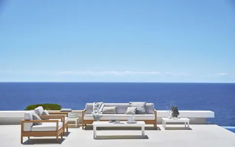Un elegante divano Bali in legno e cuscini chiari, con vista sul mare blu intenso sotto un cielo limpido.