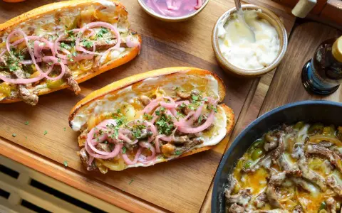 Två philly cheese steak-mackor med möra strimlor av kött, smält ost, lök och paprika, toppade med picklad rödlök och persilja.