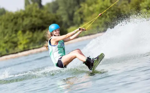 Dziewczyna w kasku i kamizelce pływa na wakeboardzie, tworząc wielki szelment wody.
