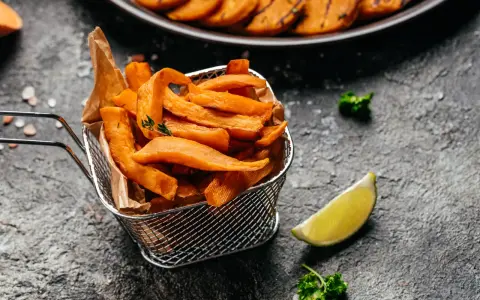 Frytki z batatów w metalowym koszyku, ozdobione gałązką tymianku, obok ćwiartki limonki i natki pietruszki.