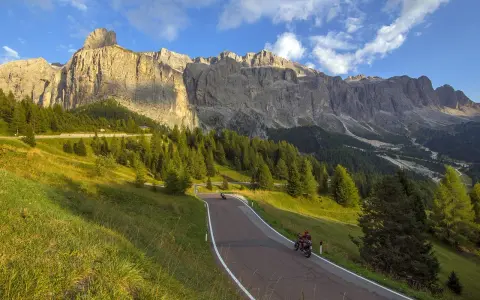 Un motociclista percorre una strada panoramica tra prati verdi e boschi di conifere, con le maestose Dolomiti sullo sfondo. Un'avventura indimenticabile in moto.