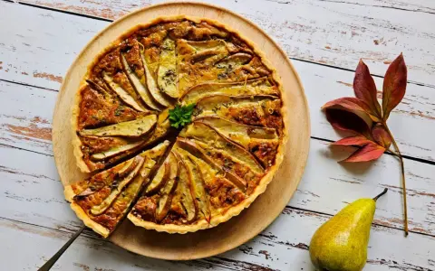 Apetyczna tarta z gruszkami, udekorowana natką pietruszki i skórką cytrynową, prezentuje się na drewnianej desce. Obok leży świeża gruszka i jesienne liście.