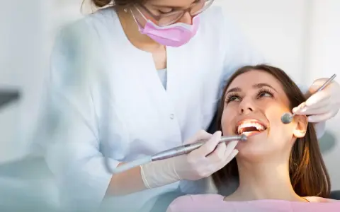 Dentystka w masce i rękawiczkach bada zęby pacjentki za pomocą lusterka i wiertła.