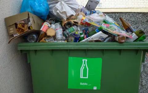 Przepełniony zielony kontener na śmieci z butelkami i szkłem, z zieloną naklejką "Szkło".