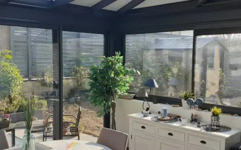 Intérieur lumineux d'une véranda triple vitrage, avec vue sur un jardin verdoyant et une commode blanche décorée.