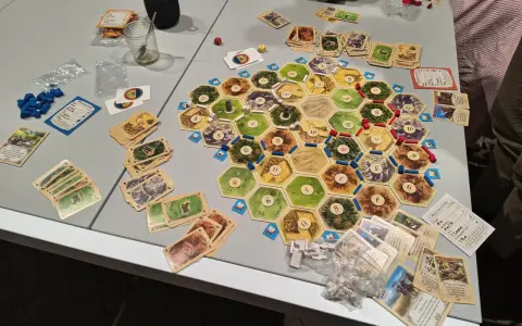 Plansza do gry Catan, karty surowców i rozwoju, pionki. Gracze zastanawiają się nad kolejnością budowania, by zdobyć jak najwięcej punktów.