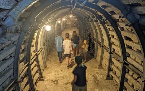 Rodzina zwiedza podziemia Sandomierz, idąc przez oświetlony tunel z kamiennymi ścianami i metalowymi wzmocnieniami.