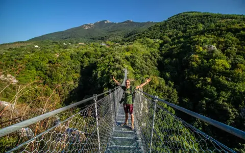 Uomo felice sul laviano ponte tibetano, con montagne verdi e cielo azzurro sullo sfondo.