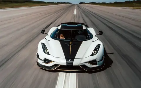 Biały Koenigsegg Regera z czarnym pasem i złotymi akcentami pędzi po pasie startowym, zostawiając za sobą smugę prędkości.