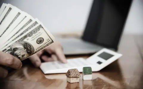 Ręka trzyma banknoty 100 USD, obok kalkulator, laptop i miniaturowe domki. Zastanawiasz się, ile wynosi zasiłek socjalny na mieszkanie?
