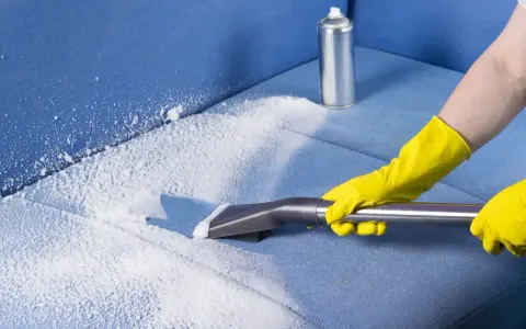 Mani con guanti gialli eseguono il lavaggio divano a secco con schiuma e aspiratore su tessuto blu.