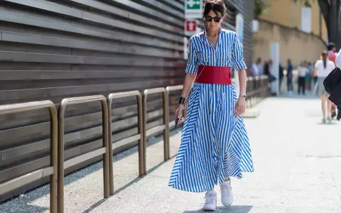 Donna con vestito a righe blu e bianche, cintura rossa e sneakers bianche cammina per strada.
