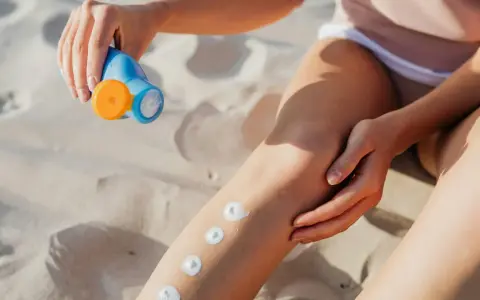 Main sur le sable, une main applique des points de crème solaire sur une jambe. Attention, cette crème solaire perimee pourrait ne plus protéger efficacement.