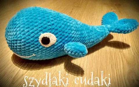 Uroczy, niebieski wieloryb na szydełku, z białym oczkiem i czarną źrenicą, leży na drewnianej podłodze.