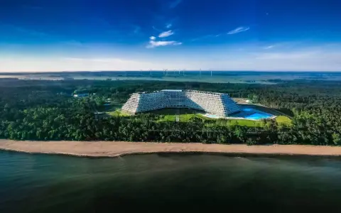 Hotel Gołębiewski w Pobierowie, otoczony lasem, z basenem i widokiem na morze. W oddali widać farmę wiatrową.