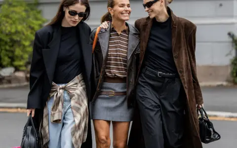 Drei Frauen in komischer Mode, die lachend nebeneinander gehen. Eine trägt einen braunen Lederblazer, die andere einen langen braunen Mantel.