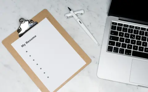 Na biurku leży laptop, notatnik z napisem "My Resume" i długopis. Czas przygotować CV, uwzględniając stopnie naukowe po angielsku.