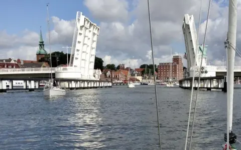 Eine Klappbrücke öffnet sich für Segelboote in Kappeln an der Schlei. Im Hintergrund die Stadt mit Kirchturm und Backsteingebäuden.