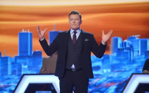 Rafał Brzozowski w garniturze, z rękami uniesionymi na boki, prowadzi program "Jaka to melodia?".