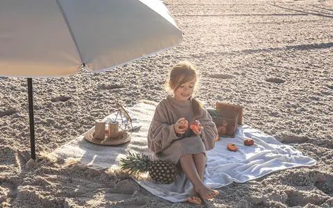 Dziewczynka na plaży pod parasolem, z owocami i ananasem. Idealny zestaw, by wiedzieć, co zabrać na wakacje nad morze z dzieckiem.