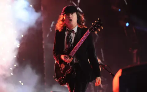 Ein Mann mit Hut und Schlagzeug spielt auf einer ACDC Gitarre, umgeben von Rauch und Lichtern.