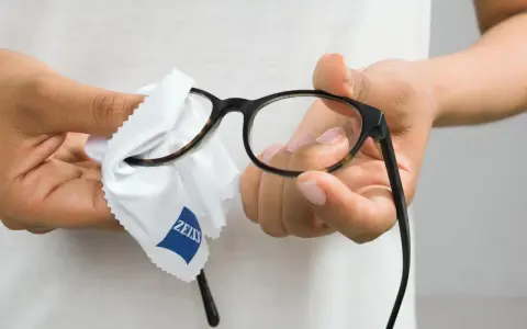 Här putsas glasögon med en mjuk trasa. En person håller i bågarna och putsar linserna med en vit trasa med Zeiss-logga.