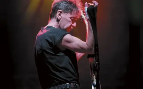 Muzyk z zespołu KSU z gitarą na scenie, w czarnej koszulce i z charakterystyczną fryzurą.