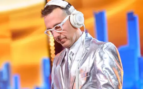 DJ w błyszczącej marynarce i białych okularach, słuchawki na uszach, gotowy do zagrania "Jaka to melodia".
