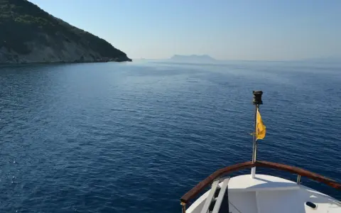 La prua di una barca naviga verso un'isola lontana, un viaggio che evoca la poesia di Itaca.