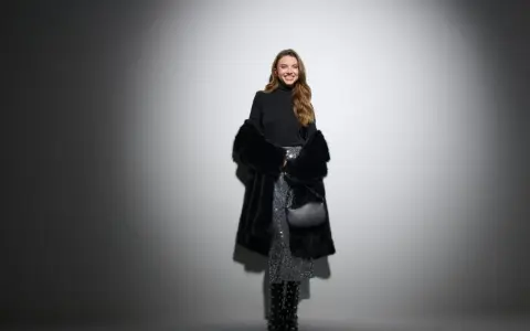 Uśmiechnięta kobieta w błyszczącej spódnicy i futrzanym płaszczu. Czy do filharmonii trzeba ubrać się elegancko? Ta stylizacja sugeruje, że tak!