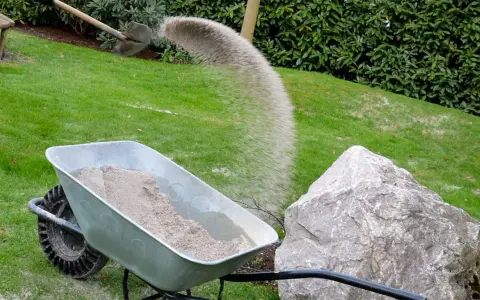 Schubkarre mit Sand, während eine Schaufel Sand auf den Rasen rieselt.
