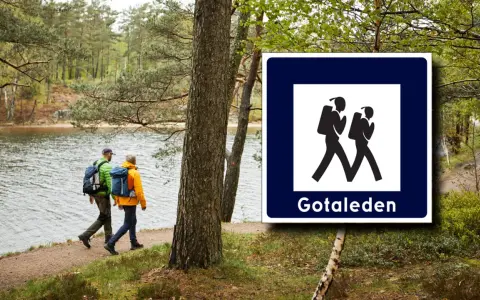 Två vandrare följer en stig vid en sjö, med en skylt för Gotaleden etapper.