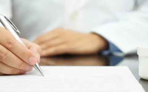 Lekarz pisze receptę farmaceutyczną, obok stoją białe opakowania leków.