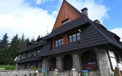 Kamienny schron turystyczny pod gorącymi szczytami, z drewnianą przybudówką i czarnym dachem, otoczony lasem świerkowym.