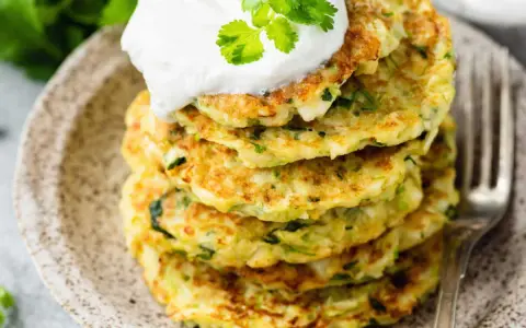 Pile de galettes de courgette dorées, garnies de crème et de coriandre fraîche, sur une assiette en céramique.
