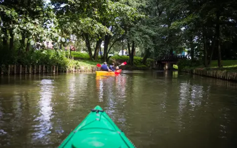 Dwie osoby płyną kajakiem po rzece, odkrywając malownicze radunia szlaki. Zielony kajak z przodu zaprasza do dalszej podróży.