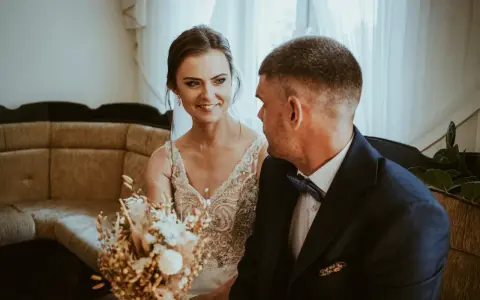 Uśmiechnięta panna młoda w sukni z koronkami i pan młody w granatowym garniturze. W tle kanapa i zasłony. Krótka ceremonia, bo ile trwa ślub cywilny?