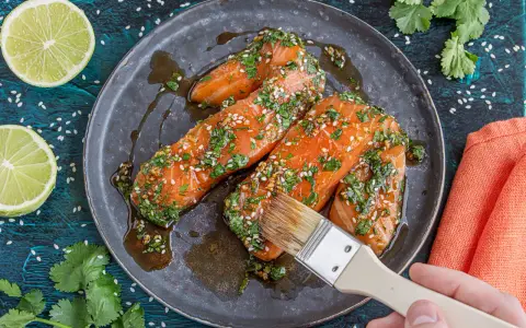 Lachsfilets werden mit einer asiatischen Marinade aus Sojasauce, Ingwer, Knoblauch, Kräutern und Sesam bestrichen. Dazu Limettenspalten und Koriander.