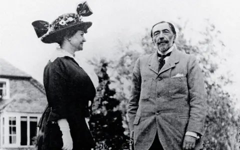 Józef Korzeniowski (aktor) w eleganckim garniturze stoi obok kobiety w ozdobnym kapeluszu.