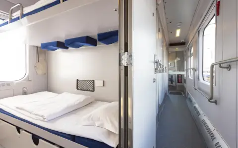 Wnętrze wagonu sypialnego z przygotowanym łóżkiem i korytarzem z drzwiami do przedziałów.