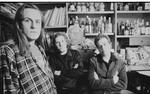Tres hombres posan en blanco y negro, con estanterías de discos y botellas detrás.