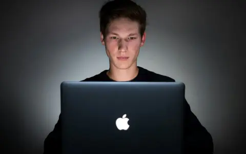 Młody mężczyzna pochylony nad laptopem, jego twarz oświetlona ekranem. Czy to początek uzależnienia od internetu?