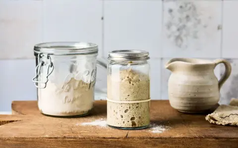 Zwei Gläser mit Sauerteig Weizen, eines mit aktivem, blasigem Sauerteig, das andere mit trockenem Ansatz. Daneben ein kleiner Krug.