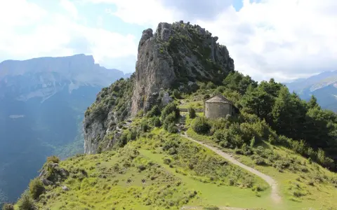 Pequeña ermita de piedra en la ruta de las ermitas, rodeada de vegetación y con imponentes montañas al fondo.
