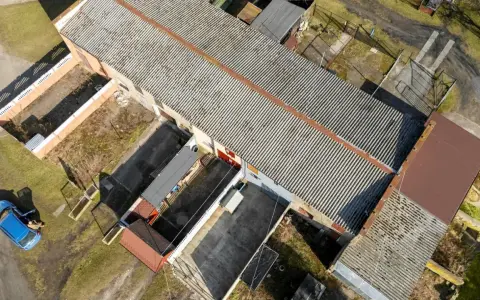 Długi budynek gospodarczy na zgłoszenie z falistym dachem, otoczony innymi mniejszymi obiektami i zielenią. Obok stoi niebieski samochód.