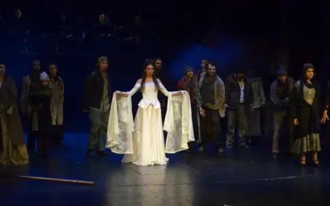 Scena z musicalu "Człowiek z La Manchy". W centrum młoda kobieta w białej sukni, otoczona przez grupę postaci w strojach z epoki.