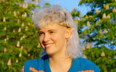 Persona sonriente con un moderno mullet rubio, aretes y un suéter azul con rayas naranjas, frente a un árbol en flor.
