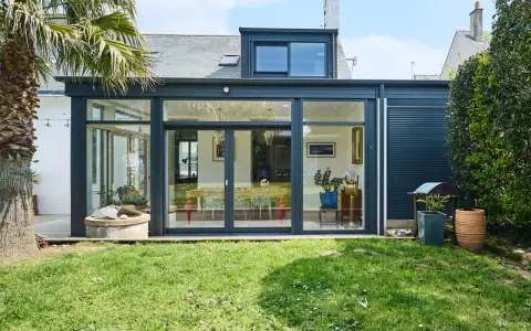Une extension de maison moderne avec une immense baie vitrée s'ouvrant sur un jardin verdoyant. Une palme se dresse à gauche.