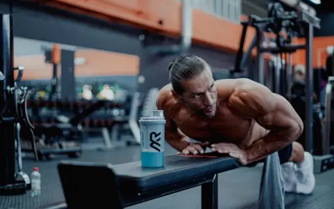 Man gör armhävningar på en bänk i gymmet, en shaker med blå dryck står bredvid. Detta är ett exempel på hur byggs muskler.
